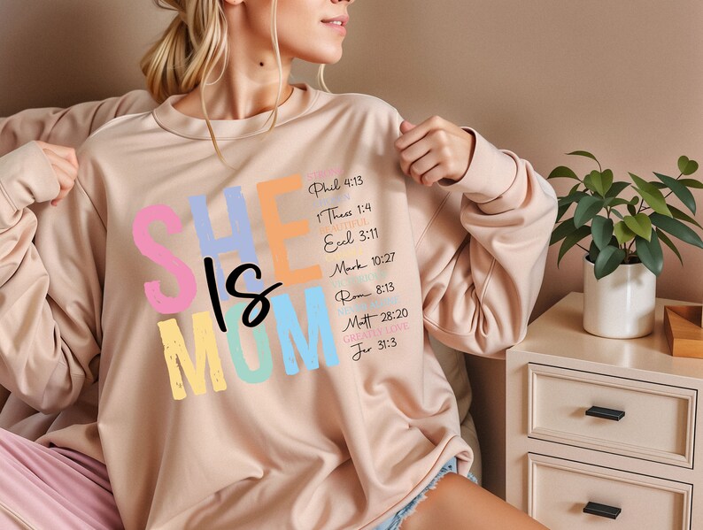 May include: A light pink sweatshirt with the text "SHE IS MOM" in colorful letters. The text is surrounded by bible verses in a smaller font.