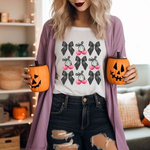 May include: A white t-shirt with a black and white cherry and bow design. The shirt is being worn by a person holding two orange pumpkin-shaped cups.