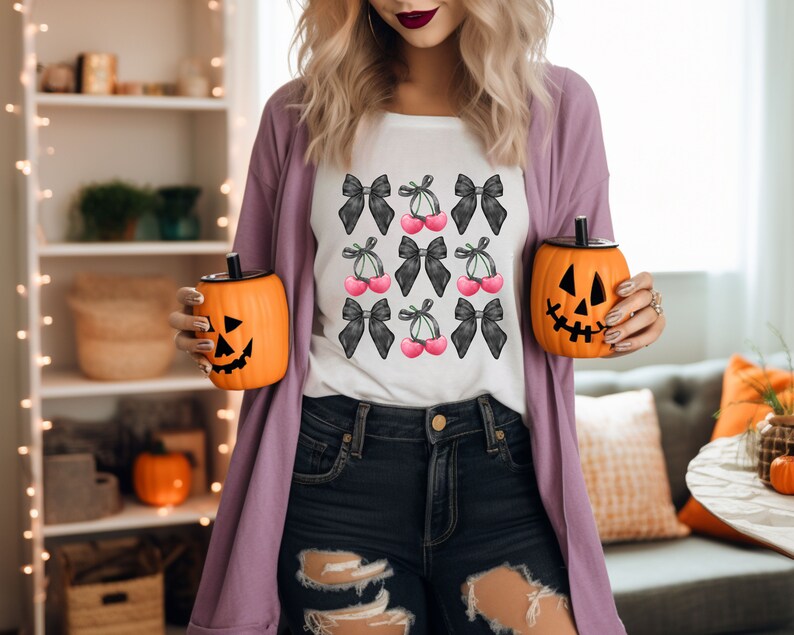 May include: A white t-shirt with a black and white cherry and bow design. The shirt is being worn by a person holding two orange pumpkin-shaped cups.