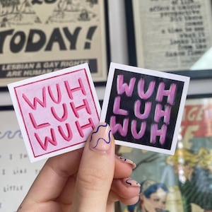 May include: Two square stickers with the text "WUH LUH WUH" in pink lettering. One sticker has a pink background, the other has a black background. The stickers are held up by a hand with painted nails.