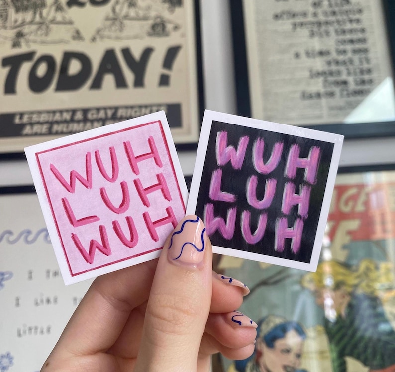 May include: Two square stickers with the text "WUH LUH WUH" in pink lettering. One sticker has a pink background, the other has a black background. The stickers are held up by a hand with painted nails.
