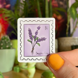 Pode incluir: Um adesivo com um buquê de flores de lavanda roxa com uma borda branca e o texto "Sapphic City" em um fundo roxo.