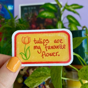 Puede incluir: Una pegatina amarilla con borde rojo y un tulipán dibujado a mano con centro amarillo y hojas verdes. La pegatina dice "los tulipes son mis flores favoritas."