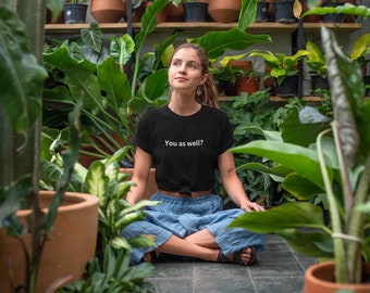 May include: A woman wearing a black t-shirt with the text "You as well?" in white lettering. She is sitting cross-legged in a lush green indoor garden setting.