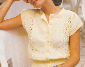 May include: A white knitted cardigan with a button-up front and short sleeves. The cardigan has a delicate lace pattern and is worn by a person sitting on a white chair. The text "DK / 8 Ply 28 - 38 ins" is visible in the top right corner of the image.