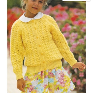 May include: A yellow knitted cardigan with a button fastening. The cardigan is designed for children and features a fine stitch pattern. The cardigan is being worn by a child with short brown hair and a floral skirt. The text on the image reads "DK / 8 Ply 20 to 30 ins 1 year to 10 years."