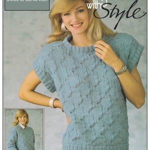 May include: A blue knitted sweater with a cable knit pattern. The sweater has a round neckline and short sleeves. The sweater is shown on a model wearing white pants. The text "Knitting with Style" is printed on the image.