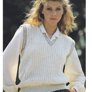May include: A white knitted vest with a V-neck and a ribbed texture. The vest has a light brown trim around the neckline and armholes. The vest is worn over a white blouse and black trousers. "MERINO DK / 8 PLY 80 TO 105cm" is written at the top of the image.