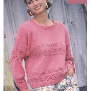 Puede incluir: Un suéter de punto rosa con un patrón de encaje. El suéter se muestra en una modelo y tiene un cuello redondo. El texto "DK / 8 Ply 3- to 40 ins" es visible en la esquina superior derecha de la imagen.