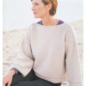 May include: A white knitted sweater with a cable knit trim around the neckline and sleeves. The sweater is worn by a person sitting on a sandy beach.
