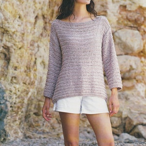 Può includere: Una donna indossa un maglione di lana marrone chiaro con scollo a barca e pantaloncini bianchi. Il maglione è realizzato con un filato a nastro e ha una vestibilità ampia. Il testo "Celeste Ribbon Yarn 32 - 42 ins" è visibile nella parte superiore dell'immagine.