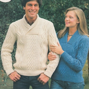 May include: A couple wearing matching cable knit jumpers. The man's jumper is cream coloured and the woman's jumper is blue. The jumpers have a v-neck and a collar that folds over. The text "Aran 32-44 ins" is in the top right corner of the image.