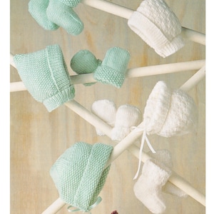 Op de afbeelding: Een set gebreide baby mutsen, bonnetjes, slofjes en wanten in wit en lichtgroen. De items hangen aan een houten rek. De tekst "Hats, Bonnets, Bootees, Mittens DK / 8 Ply 2 sizes: 0-6 months & 6-12 months" is boven de items gedrukt.