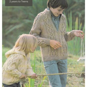 Könnte beinhalten: Zwei Kinder tragen gestrickte Aran-Pullover, einen in Beige und einen in Weiß, und stehen in einem grasbewachsenen Bereich. Der beige Pullover hat ein Zopfmuster und der weiße Pullover eine Kapuze. Der Text "Aran/Worsted Jacket" ist oben im Bild sichtbar. Der Text "22 to 32 inch chest" und "2 years to Teens" ist ebenfalls sichtbar.