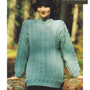 May include: A teal blue cable knit jumper with a round neckline and long sleeves. The jumper has a leaf-shaped design at the bottom hem. The model is wearing a black hat and is standing in front of a tree trunk. DK/8 Ply. 3 sizes: 30-32 to 38-40 ins.