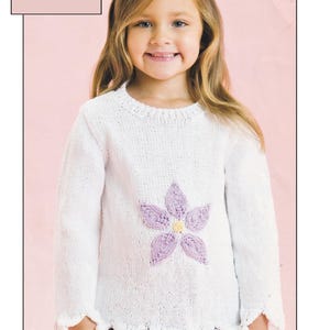 Puede incluir: Suéter blanco de punto con un diseño de flor morada para un niño. El suéter tiene un dobladillo festoneado y pompones en las mangas. Algodón DK / 8 Ply. De 0 a 4 años. 16 a 24 pulgadas de pecho.