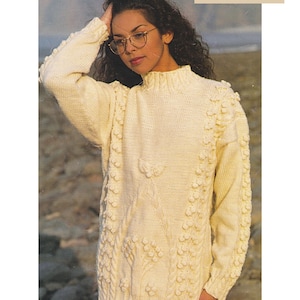 Peut inclure: Un pull en maille aran blanc à col montant et motif en tricot torsadé. Le pull a un design texturé avec de petites boules et est présenté sur un mannequin sur fond de plage. Aran / Worsted Yarn 32-42 pouces