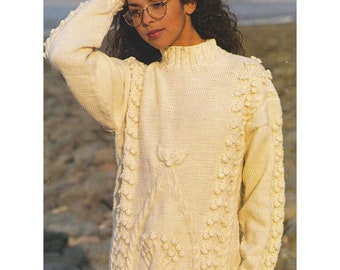 May include: A white aran knit sweater with a high neck and a cable knit pattern. The sweater has a textured design with small bobbles and is shown on a model against a beach background. Aran / Worsted Yarn 32-42 ins