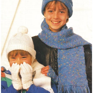 Könnte beinhalten: Zwei Kinder tragen gestrickte Winteraccessoires. Das Mädchen links trägt eine weiße Mütze mit Bommel, weiße Fingerlose Handschuhe und einen weißen Schal. Das Mädchen rechts trägt eine blaue Mütze mit weißem Bommel, einen blauen Schal mit weißen und rosa Punkten und blaue Fingerlose Handschuhe. Der Text "DK Yarn 4-13 years" ist im Bild sichtbar.
