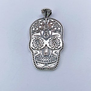 May include: Silver pendant in the shape of a sugar skull with intricate details. The skull has a floral design around the eyes and a detailed mouth.