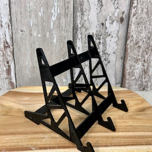 Könnte beinhalten: Schwarzer Acryl-Präsentationsständer mit geometrischem Design. Der Ständer besteht aus zwei Teilen, die miteinander verbunden sind. Der Ständer ist so konzipiert, dass er eine Vielzahl von Gegenständen aufnehmen kann, wie z. B. Bücher, Fotos oder kleine Skulpturen.