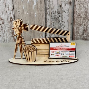 May include: A wooden movie-themed decorative piece featuring a film camera on a tripod, a clapperboard, and a popcorn container. Includes a gift card holder with the text "WUNSCHGUTSCHEIN" and the names "Manu & Mike" on the base.