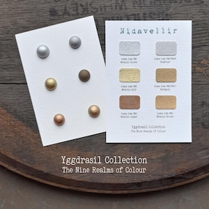 Könnte beinhalten: Ein Set aus sechs kugelförmigen Knöpfen in Silber-, Gold- und Kupfertönen auf einer weißen Karte, neben einer Referenzkarte mit der Aufschrift Nidavellir Yggdrasil Collection The Nine Realms of Colour, die sechs rechteckige Farbmuster zeigt.