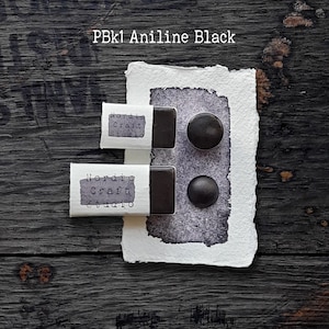 Könnte beinhalten: Zwei rechteckige Näpfchen mit PBk1 Aniline Black Aquarellfarbe mit weißen Papieretiketten, auf denen Nordic Craft Studio steht, sowie zwei kreisförmige schwarze Farbproben auf einem strukturierten weißen Papier mit einem dunkelgrauen Farbfleck.