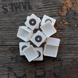 Puede incluir: Un conjunto de piezas cuadradas de cerámica blanca con un círculo gris oscuro en el centro y piezas rectangulares blancas a juego. Los objetos están dispuestos sobre una superficie de madera oscura y texturizada.