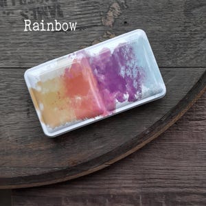 Könnte beinhalten: Ein rechteckiger Aquarellfarbkasten aus weißem Metall mit einem abstrakten, regenbogenfarbenen Farbauftrag auf dem Deckel. Das Wort Rainbow ist in weißer Schrift oben links auf den Deckel gedruckt und zeigt gelbe, orange, violette und blaue Farbtöne.