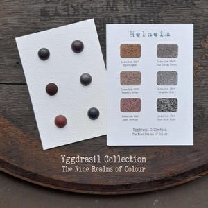 Könnte beinhalten: Eine weiße Karte mit sechs runden Knöpfen in Braun- und Grautönen sowie eine zweite weiße Karte mit dem Titel Helheim, die sechs rechteckige Farbmuster mit Indexcodes und Namen wie Burnt Umber und Graphite Grey zeigt.