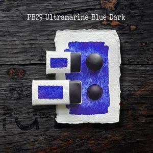 Könnte beinhalten: Ein rechteckiges Stück weißes, strukturiertes Papier mit einem großen ultramarinblauen Farbfleck, zwei schwarzen runden Farbnäpfchen und zwei weißen Papierhüllen mit der Aufschrift PB29 Ultramarine Blue Dark und blauen Farbproben.