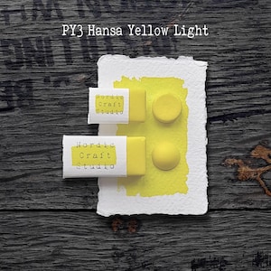 May include: A close-up of art supplies, including two rectangular paint sticks and two round paint samples in a vibrant yellow hue, labeled "PY3 Hansa Yellow Light." The paint is on a textured white paper, with the text "Nordic Craft Studio" on the paint sticks.