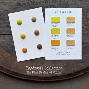 Könnte beinhalten: Zwei weiße Karten mit der Yggdrasil Collection The Nine Realms of Colour. Eine Karte zeigt sechs kreisförmige Knöpfe in Gelb-, Orange- und Brauntönen. Die andere Karte zeigt sechs rechteckige Farbmuster mit entsprechenden Beschriftungen.