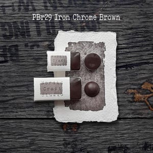 May include: Brown watercolour paint samples in square and round shapes on textured white paper. The text "PBr29 Iron Chrome Brown" is at the top. The paper is set on a dark wood surface. The labels read "Nordic Craft Studio".