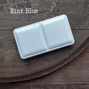 Pode incluir: Uma godé retangular para aguarela na cor azul menta com dois compartimentos quadrados distintos e rebaixados. A superfície do recipiente de plástico é lisa e brilhante, apresentando um design limpo e simples para conter pastilhas de pigmento.