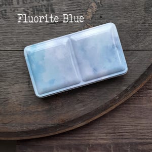 Könnte beinhalten: Eine rechteckige Metalldose mit Klappdeckel und einem aquarellartigen Muster in Hellblau und Weiß. Der Text Fluorite Blue ist in Weiß auf der oberen linken Seite des Deckels aufgedruckt, der zwei deutlich vertiefte Bereiche aufweist.