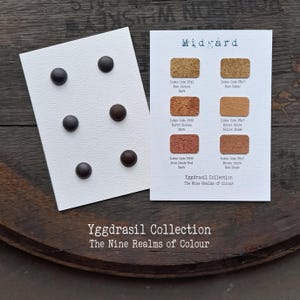 Könnte beinhalten: Eine weiße Karte mit sechs dunklen runden Knöpfen und eine zweite weiße Karte mit dem Titel Midgard Yggdrasil Collection The Nine Realms of Colour mit sechs rechteckigen Farbmustern, beschriftet mit Indexcodes und Farbnamen.