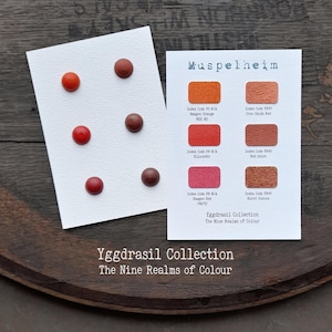 Könnte beinhalten: Zwei weiße Karten aus der Yggdrasil Collection The Nine Realms of Colour. Eine Karte zeigt sechs runde Farbmuster in Rottönen. Die andere Karte enthält sechs rechteckige Farbfelder mit Beschriftungen wie Iron Oxide Red und Burnt Sienna.