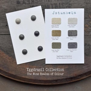 Könnte beinhalten: Zwei weiße Karten mit der Jotunheim Yggdrasil Kollektion. Eine Karte zeigt sechs kreisförmige Knöpfe in Grau- und Anthrazittönen. Die andere Karte enthält sechs rechteckige Farbmuster mit entsprechenden Farbcodes und Bezeichnungen.