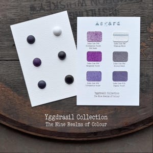 Könnte beinhalten: Zwei weiße Karten der Asgard Yggdrasil Kollektion. Eine Karte zeigt sechs kreisförmige Farbmuster in Weiß-, Violett- und Schwarztönen. Die zweite Karte enthält sechs rechteckige Farbfelder mit Beschriftungen für Farbnamen und Index-Codes.
