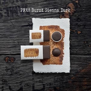 Könnte beinhalten: Zwei rechteckige Blöcke und zwei runde Näpfchen mit dunkelbrauner Aquarellfarbe, beschriftet mit PR101 Burnt Sienna Dark. Die Farbe liegt auf einem strukturierten weißen Papier mit einem braunen Farbfleck und dem Nordic Craft Studio Logo.