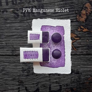 Könnte beinhalten: Zwei rechteckige und zwei runde Näpfchen mit PV16 Manganese Violet Aquarellfarbe. Die Näpfchen sind in weiße Papieretiketten mit dem Text Nordic Craft Studio eingewickelt. Ein weißes, strukturiertes Papier zeigt einen violetten Farbauftrag.