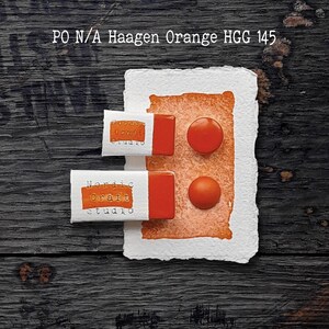 Könnte beinhalten: Zwei rechteckige orangefarbene Farbnäpfchen mit der Aufschrift Nordic Craft Studio und zwei runde orangefarbene Farbtropfen auf einem strukturierten weißen Papiermuster. Der Text PO N/A Haagen Orange HGG 145 steht oben im Bild.