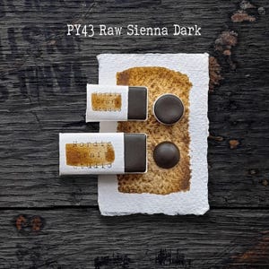 Könnte beinhalten: Zwei rechteckige Aquarellnäpfchen und zwei runde Aquarellnäpfchen in der Farbe PY43 Raw Sienna Dark. Die Näpfchen sind mit weißen Papierbanderolen versehen, die den Text Nordic Craft Studio tragen. Ein Farbmuster befindet sich auf strukturiertem Papier.