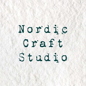 Puede incluir: Un fondo de papel blanco texturizado con el texto verde oscuro Nordic Craft Studio impreso en una fuente estilo m&aacute;quina de escribir. Las letras tienen un aspecto desgastado y estampado, creando un alto contraste sobre la superficie rugosa.