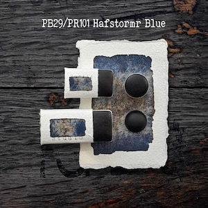 May include: Two rectangular black watercolour pans and two circular black watercolour pans are arranged on a piece of textured white paper with a blue and brown painted swatch. The text PB29/PR101 Hafstormr Blue is visible at the top.