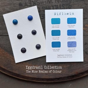 Könnte beinhalten: Eine weiße Karte mit sechs blauen und schwarzen runden Knöpfen und eine zweite weiße Karte mit dem Titel Niflheim aus der Yggdrasil Collection, die sechs blaue Farbmuster mit Indexcodes und Namen wie Cobalt Cerulean und Ultramarine zeigt.