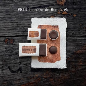 Puede incluir: Muestras de pintura de acuarela Iron Oxide Red Dark. La imagen muestra dos bloques de pintura rectangulares en envoltorios de papel blanco, dos muestras de pintura redondas y dos bloques de pintura rectangulares. El texto "PR101 Iron Oxide Red Dark" está en la parte superior.