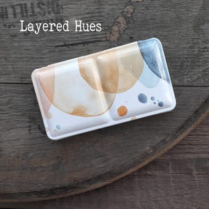 May include: A rectangular metal watercolour paint tin featuring a white lid decorated with abstract watercolour washes in shades of tan, peach, and blue. The text Layered Hues is printed in white on the upper left corner of the lid surface.